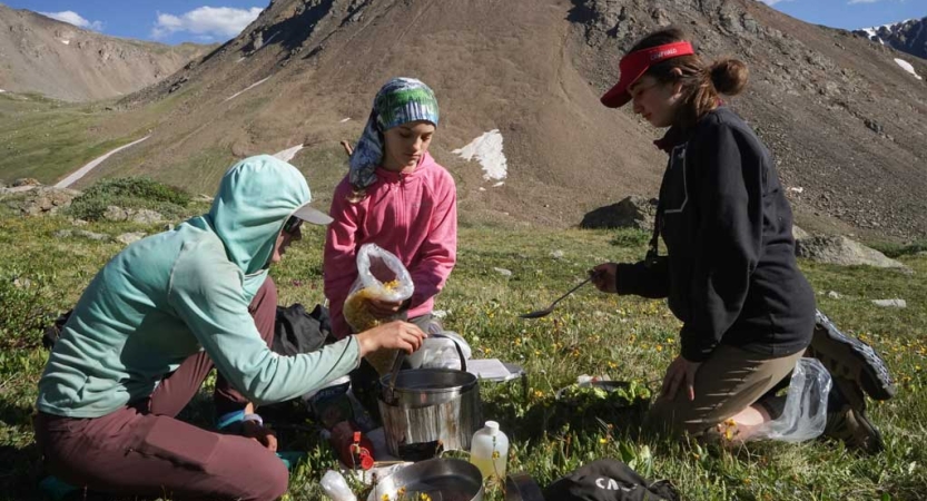 Three people gather around a cooking pot in an open space with a mountain in the background 
